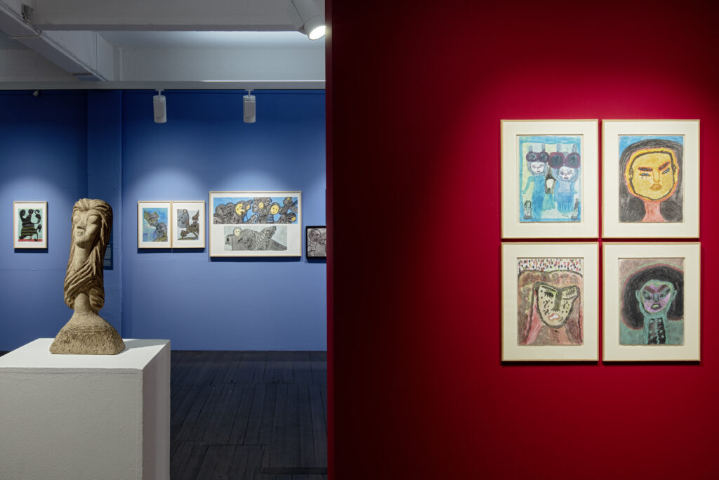 Blick in die Ausstellung mit einer Stellwand im Vordergrund der Fotografie. Im Mittelgrund steht ein Objekt auf einem Sockel. Der Hintergrund ist eine blaue Wand mit enggehängten Kunstwerken.
